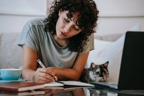 Person writing in a notebook next to a laptop and a cat