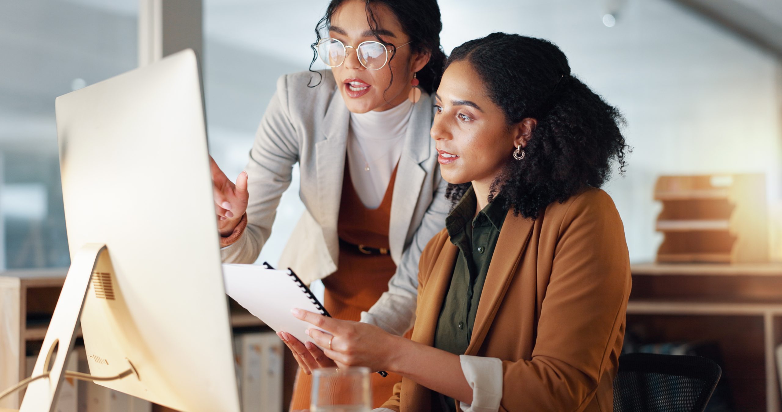 Photo of two women looking at a laptop, one woman coaching the other woman holding a piece of paper.