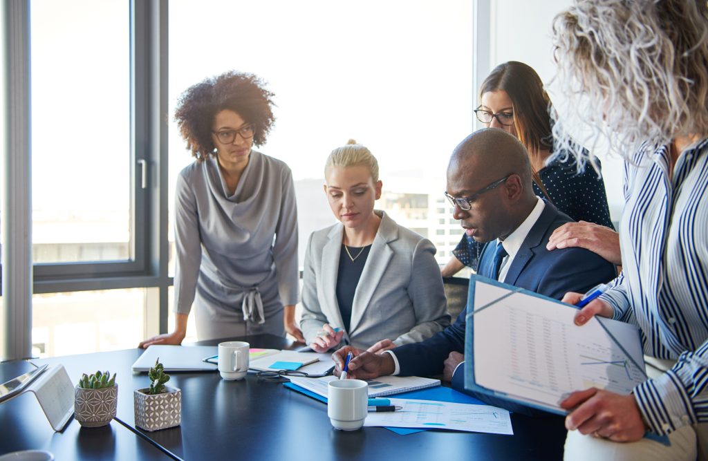 Group of focused businesspeople standing around their manager sitting at an office boardroom table reviewing charts and paperwork
